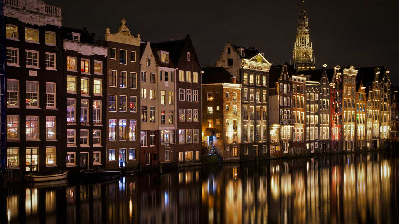 La imagen muestra las clásicas casas estrechas y altas de Ámsterdam alineadas junto a un canal, iluminadas cálidamente durante la noche. Las luces de las ventanas y fachadas se reflejan en el agua tranquila del canal, creando una vista pintoresca y encantadora de la ciudad. Al fondo, se puede ver la torre de una iglesia, añadiendo un toque histórico al paisaje urbano nocturno.