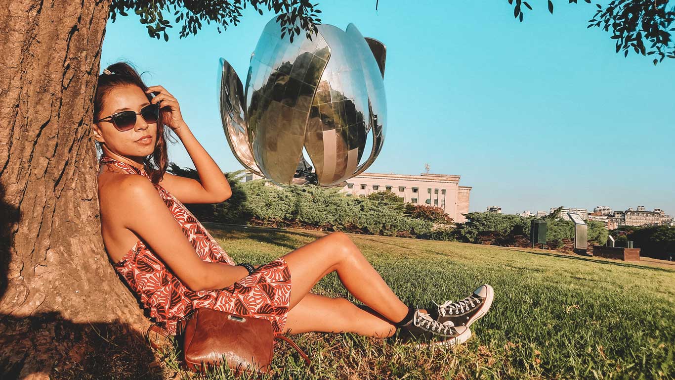 La imagen muestra a una mujer sentada bajo la sombra de un árbol, apoyada en su tronco, usando gafas de sol y un vestido veraniego con estampado. En el fondo, se observa la famosa escultura "Floralis Genérica" de metal brillante en Buenos Aires, Argentina, uno de los mejores destinos para viajar con amigos.