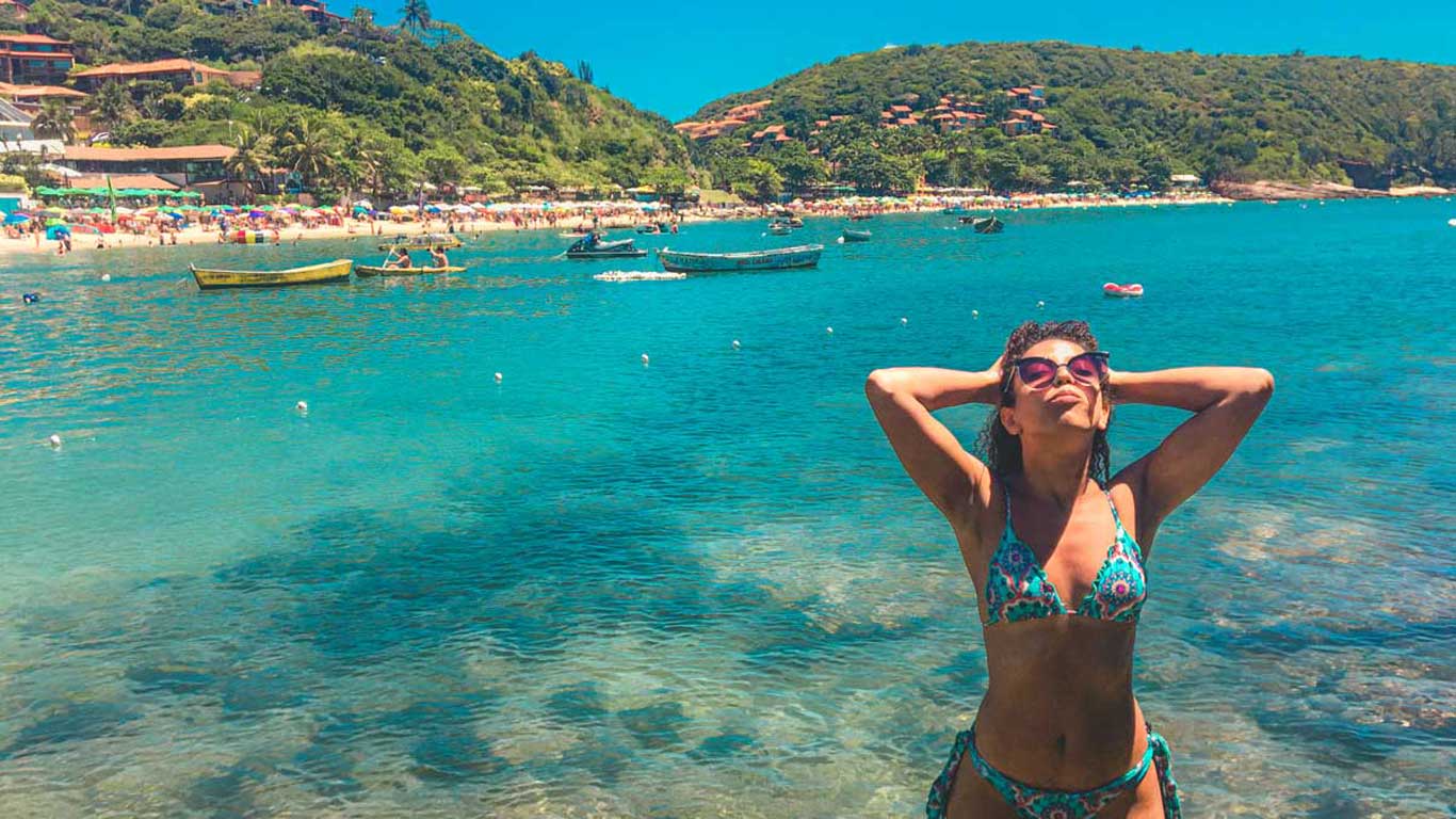 La imagen muestra a una mujer en bikini disfrutando de una playa de aguas cristalinas en Búzios, Río de Janeiro, Brasil. Al fondo, se ve una playa concurrida con sombrillas de colores, lanchas en el agua y colinas verdes cubiertas de vegetación. El día es soleado, lo que resalta los tonos azules del mar y el ambiente relajado de este destino turístico.