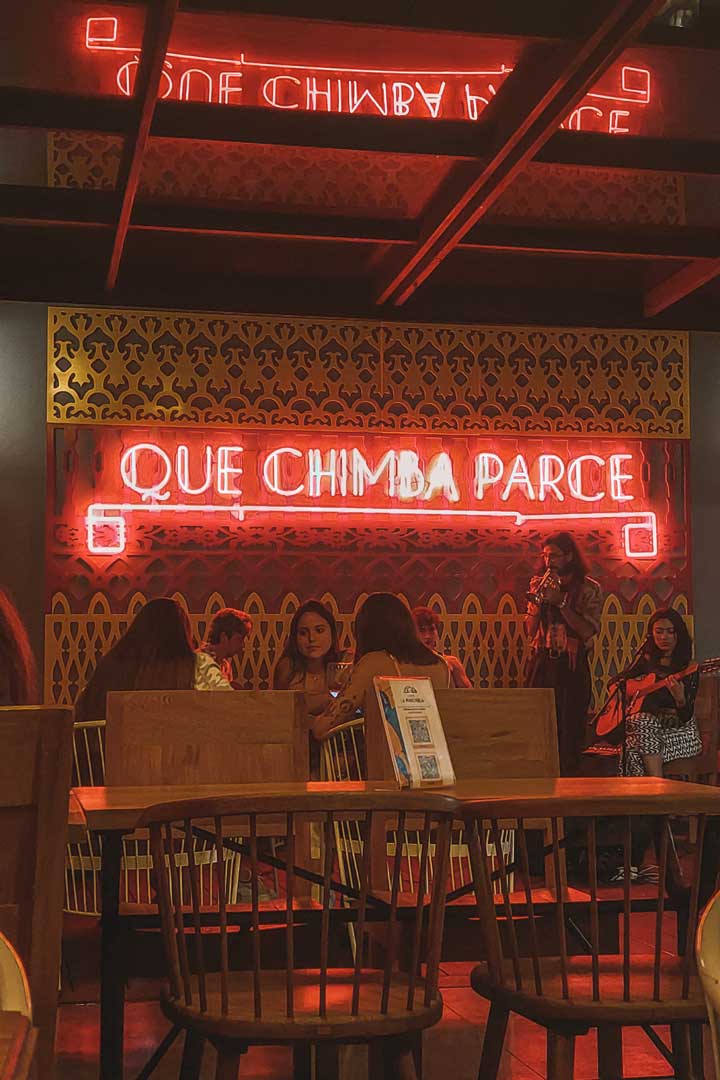 cafe-la--machuria-medellin Interior cálido de una cafetería con muebles de madera y un letrero de neón que dice “QUE CHIMBA PARCE”. Un grupo de amigos conversa mientras al fondo dos mujeres tocan música en vivo con guitarra y flauta.