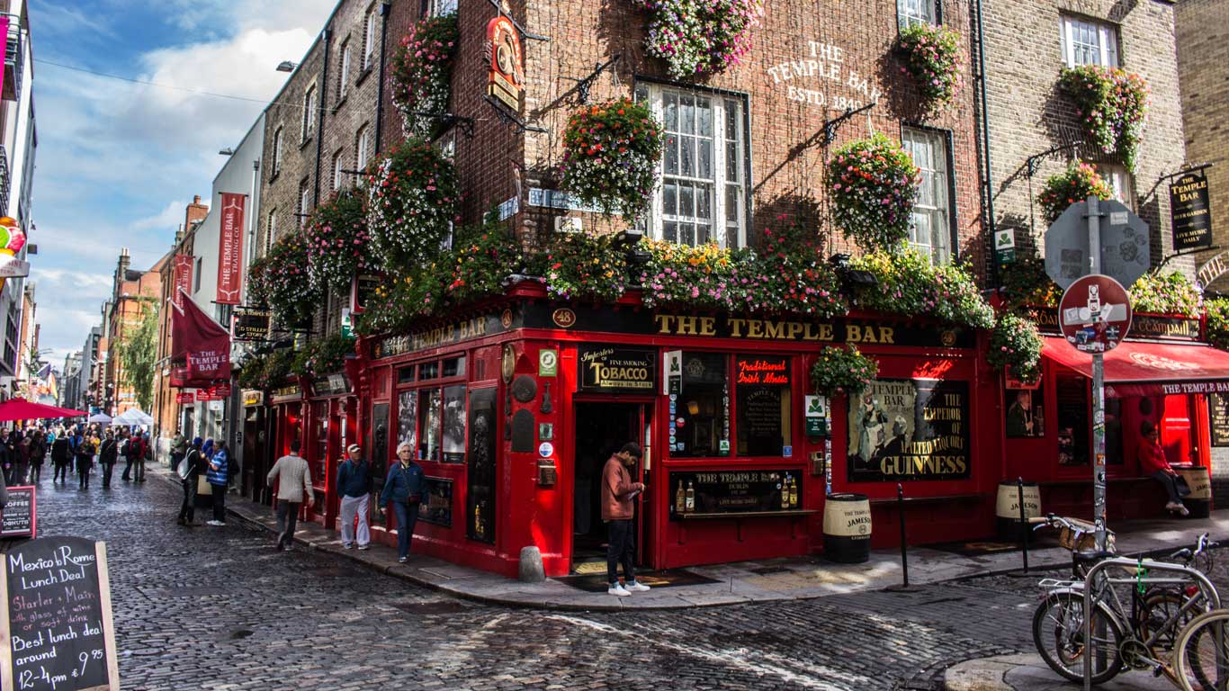 La imagen muestra la icónica fachada roja de "The Temple Bar" en Dublín, Irlanda, adornada con macetas llenas de flores coloridas. El bar está ubicado en una calle adoquinada, rodeado de personas que pasean y disfrutan del ambiente animado.