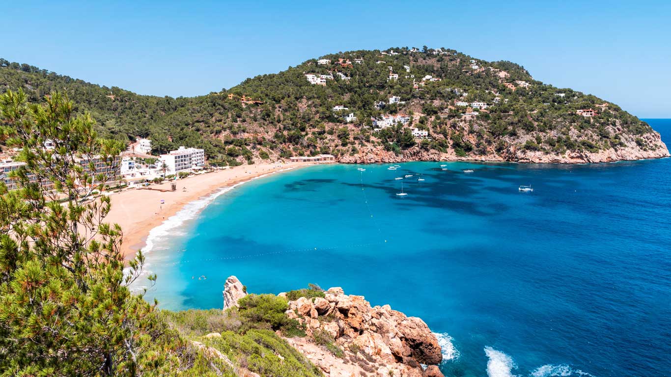 La imagen muestra una cala de aguas cristalinas en Ibiza, España, rodeada de colinas verdes con casas blancas dispersas. La playa de arena dorada se extiende a lo largo de la bahía, mientras algunas embarcaciones flotan en el mar azul turquesa. Ibiza también es uno de los mejores destinos para viajar con amigos.