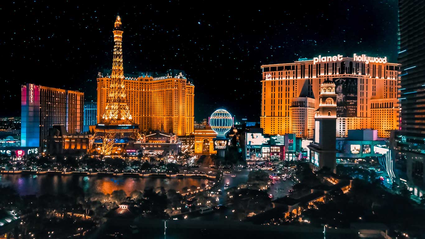 La imagen muestra una vista nocturna icónica de Las Vegas, con sus brillantes luces y famosos hoteles. Destacan la réplica de la Torre Eiffel del Paris Las Vegas, el hotel Planet Hollywood, y varias otras estructuras iluminadas que capturan la energía vibrante de la ciudad. En primer plano se puede ver parte de la fuente del Bellagio, mientras que el cielo estrellado añade un toque de magia a la escena.