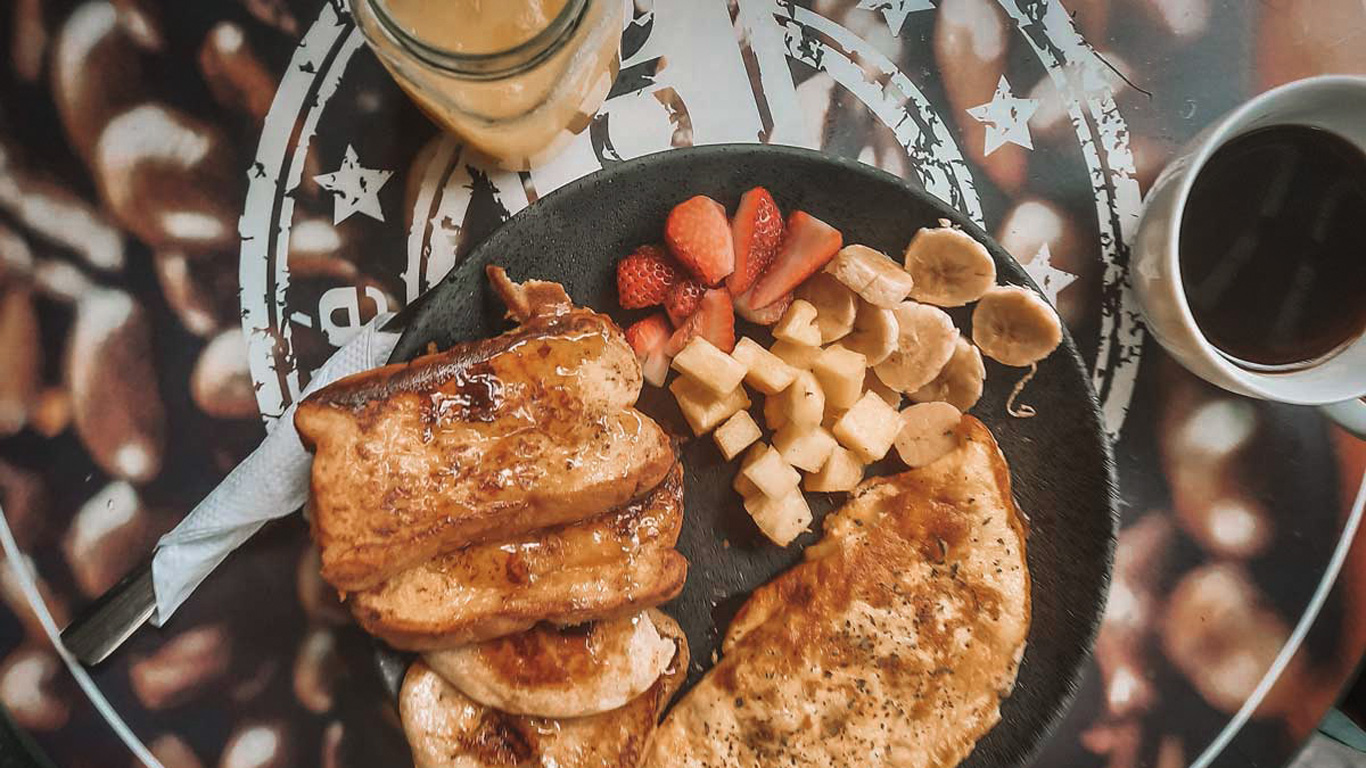 Plato de desayuno con tostadas francesas doradas con jarabe, una tortilla esponjosa y una mezcla colorida de frutas: fresas, banano y cubos de manzana. Acompañado de un café negro y un café frío en frasco de vidrio, sobre una mesa decorada con granos de café.
