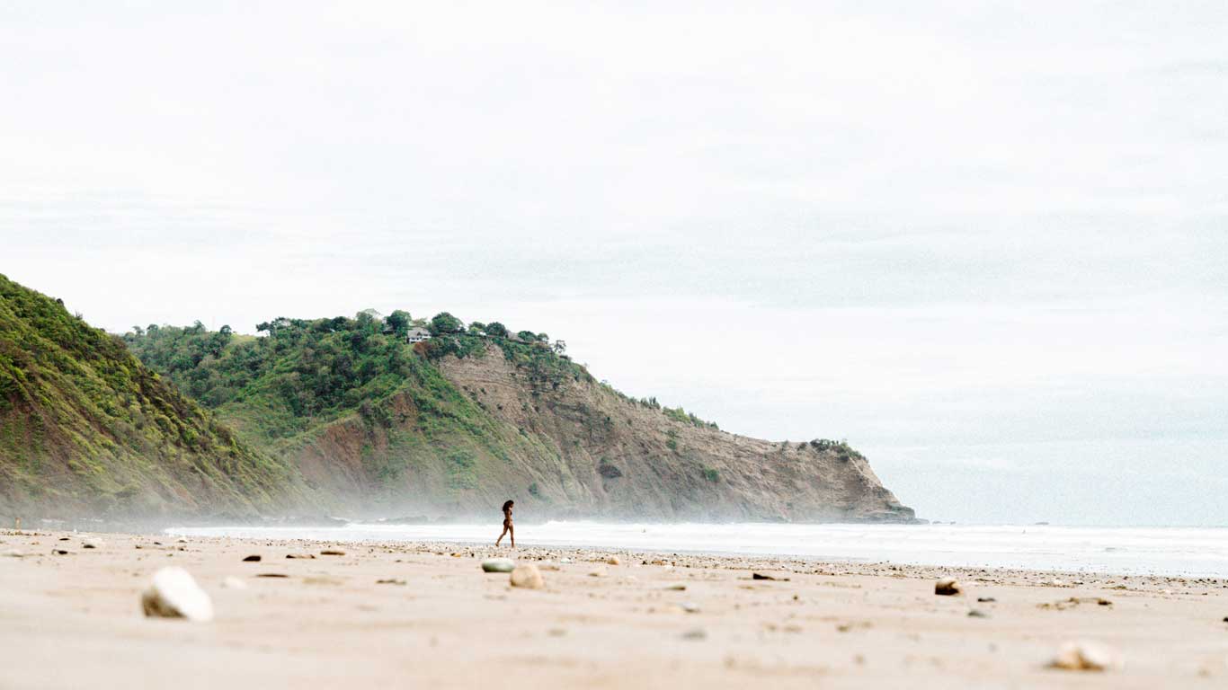 La imagen muestra una amplia playa vacía con una persona caminando por la orilla en solitario. Al fondo, se aprecian imponentes acantilados verdes que bordean la costa, bajo un cielo nublado. La escena transmite una sensación de tranquilidad en un entorno natural cerca de Montañita, Ecuador.