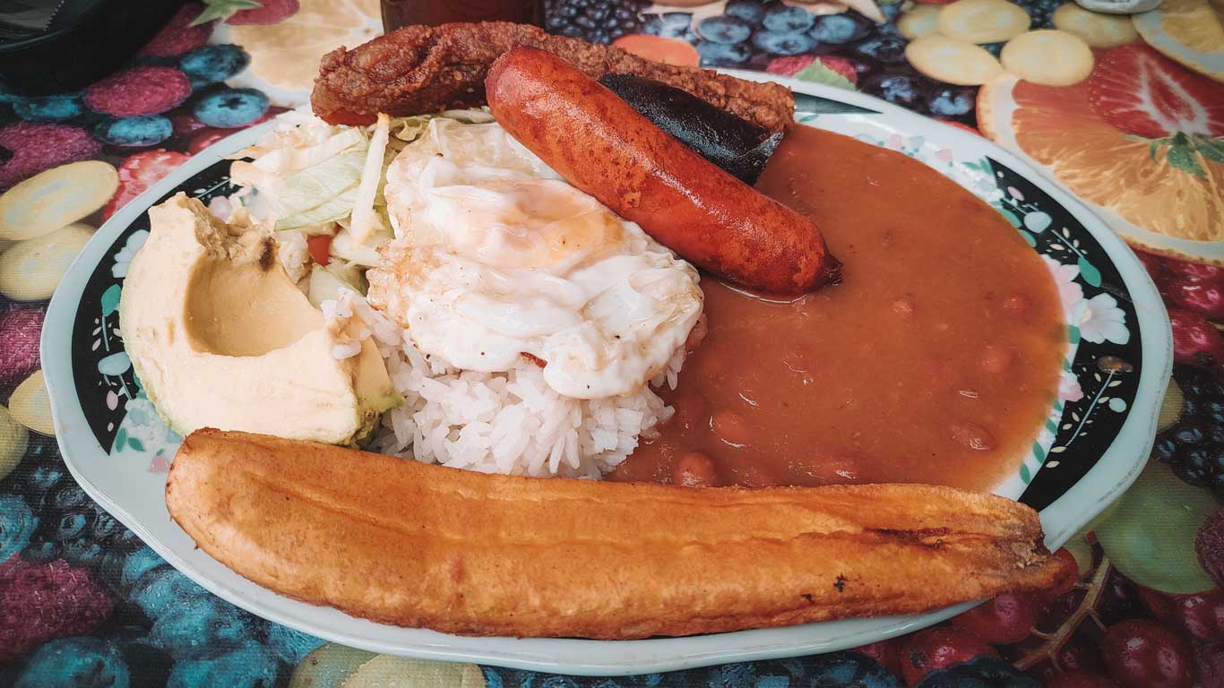 Un plato tradicional colombiano que incluye arroz blanco con un huevo frito encima, chorizo, chicharrón, tajada de plátano maduro, aguacate, ensalada y frijoles rojos. La presentación refleja la abundancia y variedad típica de la gastronomía local. El fondo colorido de la mesa complementa la vivacidad del plato.