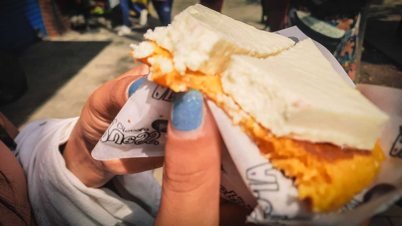 La imagen muestra una persona sosteniendo una arepa típica colombiana cubierta con una generosa porción de queso fresco. La arepa tiene un color dorado y textura esponjosa, mientras que el queso aporta un contraste suave y cremoso. Este snack representa un ícono de la gastronomía tradicional de Medellín, disfrutado comúnmente en las calles o mercados locales.