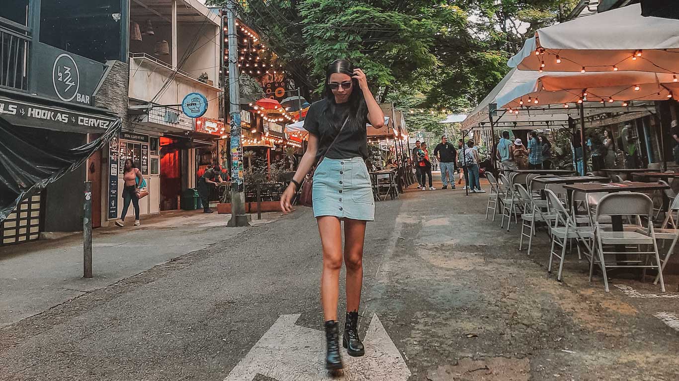 Una mujer camina por una calle peatonal en un barrio animado, rodeada de bares y restaurantes iluminados con luces cálidas. Las mesas al aire libre están decoradas con sombrillas, y al fondo se observan otras personas disfrutando del ambiente relajado. La escena refleja un entorno vibrante y moderno, ideal para el ocio y la vida nocturna.