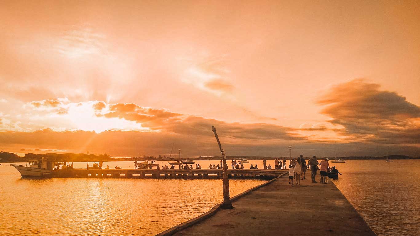 Una imagen de un muelle que se adentra en el agua al atardecer, con el cielo teñido de tonos anaranjados y dorados. Hay personas paseando y observando el paisaje, mientras un barco está atracado cerca del muelle, capturando un ambiente tranquilo y sereno.