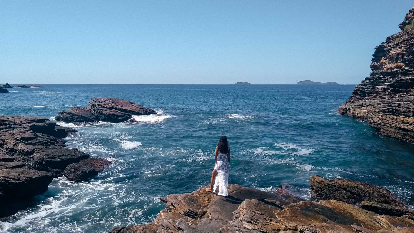 La imagen muestra a una mujer de espaldas, vestida con un traje blanco, de pie sobre unas rocas frente al mar en Búzios, Brasil. El paisaje combina aguas cristalinas de azul intenso, formaciones rocosas y un horizonte despejado con pequeñas islas en la distancia. La escena evoca tranquilidad y conexión con la naturaleza.