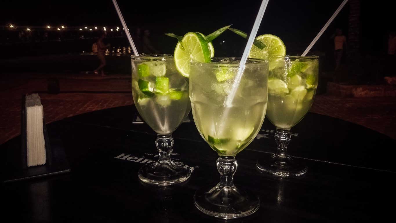 Tres copas de cócteles servidas en una mesa negra del bar Zapata, en Búzios, decoradas con rodajas de lima y trozos de lima fresca. La escena parece estar en un ambiente nocturno al aire libre, con luces difusas y algunas personas caminando en el fondo, añadiendo un aire relajado y festivo. El bar es una excelente elección de qué hacer en Búzios por la noche.