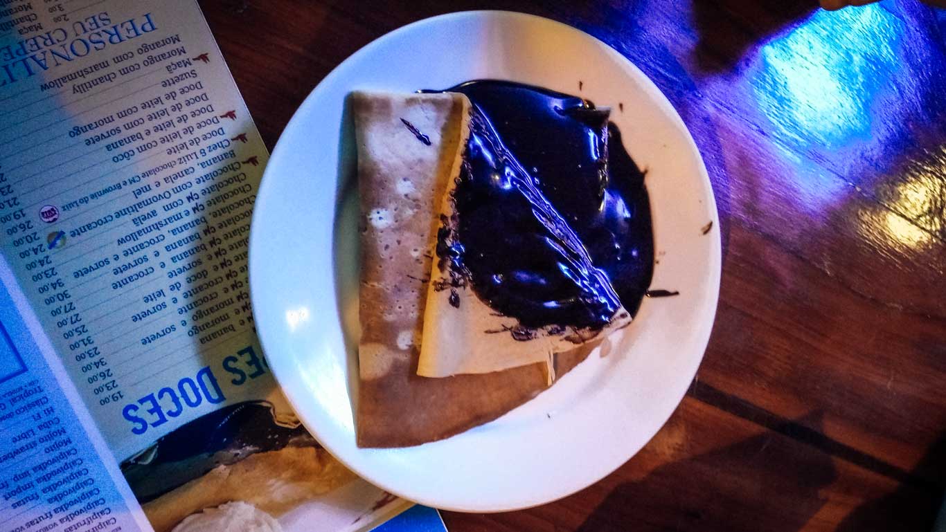 Un plato con un crepe servido en un restaurante, cubierto con abundante salsa de chocolate brillante. Al fondo, se puede observar parte de un menú con opciones de postres, todo colocado sobre una mesa de madera con iluminación cálida.