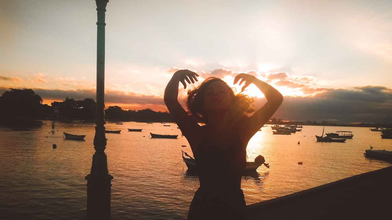 Una silueta de una mujer con los brazos levantados frente a un atardecer deslumbrante en la costa, con el cielo teñido de tonos cálidos y barcos flotando en el agua. Una farola antigua se alza a un lado, añadiendo un toque nostálgico al paisaje. La escena evoca tranquilidad y conexión con la naturaleza.