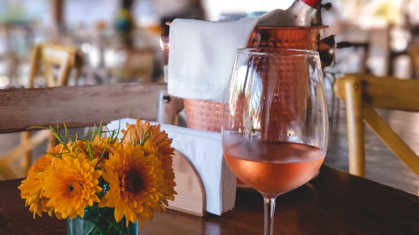 Una mesa de restaurante con un ambiente acogedor, decorada con un vaso de vino rosado, un jarrón con flores amarillas brillantes y un enfriador de cobre con una botella de vino cubierta con una servilleta blanca. El fondo desenfocado muestra un espacio relajado con sillas y mesas de madera, resaltando un ambiente casual y elegante.