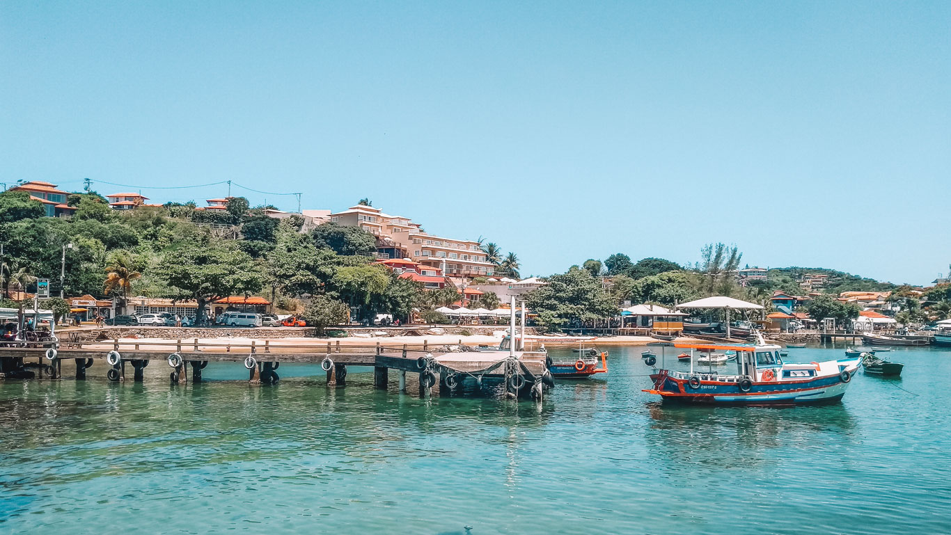 Imagen de un paisaje costero de Búzios, Brasil, con barcos de pesca anclados en un muelle de madera sobre aguas cristalinas. Al fondo, una colina cubierta de vegetación y construcciones residenciales de diferentes tamaños y colores, con un cielo azul claro arriba. La atmósfera es tranquila, destacando la belleza natural y el encanto rústico de la ciudad.