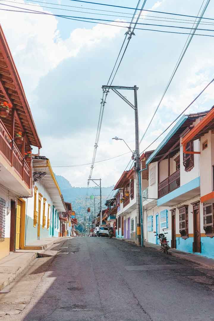 callesjardin Calle tranquila y colorida en Jardín, Colombia, flanqueada por casas tradicionales con balcones de madera, flores colgantes y montañas al fondo bajo un cielo parcialmente nublado. El estilo pintoresco del pueblo refleja su encanto típico antioqueño.