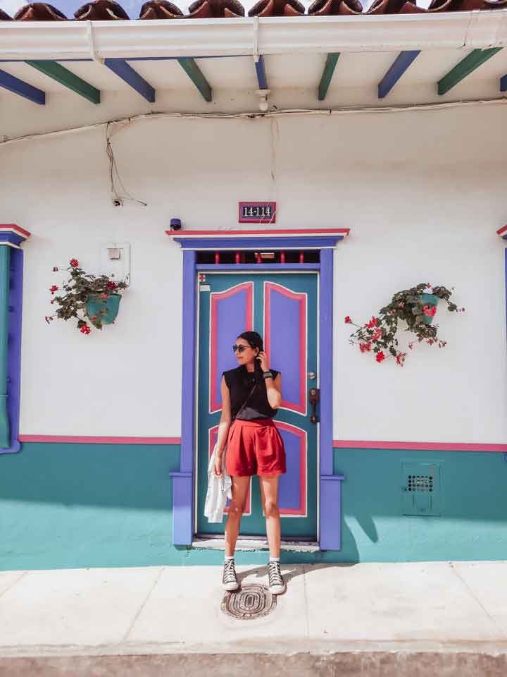 jardincolombia1 Mujer posando frente a una colorida fachada típica de Jardín, Colombia, con una puerta y ventana en tonos vivos de azul, rosa y turquesa, acompañada de macetas colgantes con flores. El estilo arquitectónico refleja la identidad vibrante y única de este encantador pueblo antioqueño.