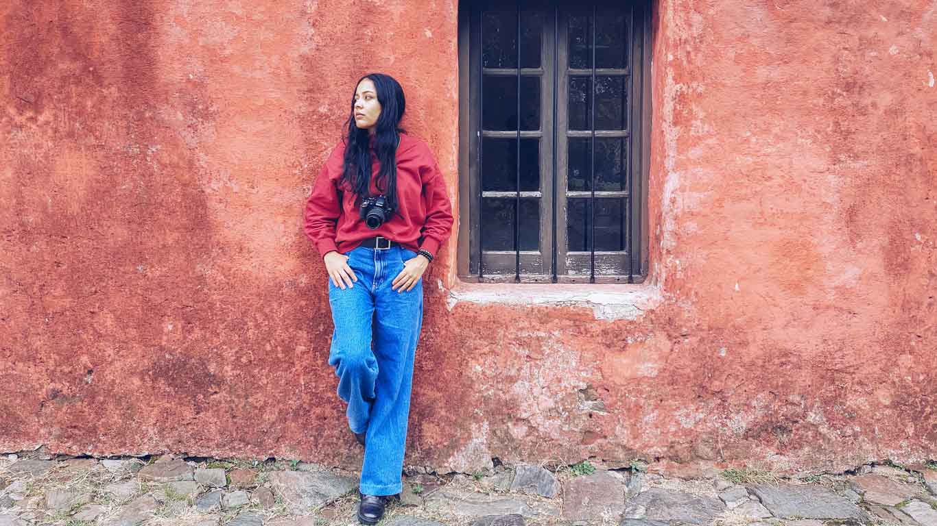 Mujer apoyada en una pared roja, al lado de una ventana, en el Casco Histórico de Colonia del Sacramento.