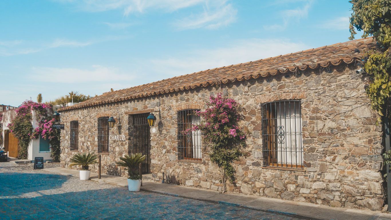 La foto muestra el Charco Hotel, un lugar acogedor con su pared de piedra adornada con algunas flores, situado en una tranquila calle pavimentada.