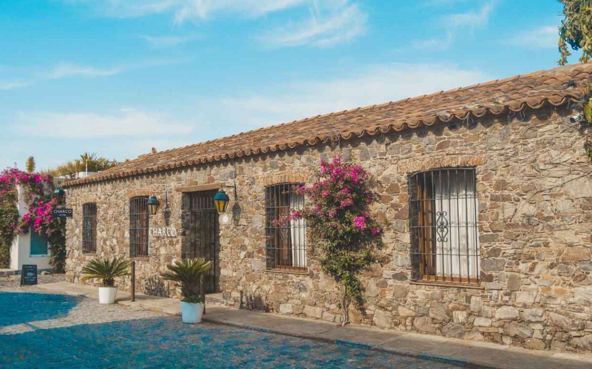 Imagen de un rústico y pintoresco hotel llamado "Charco Hotel" en Colonia del Sacramento, Uruguay. La fachada de piedra del edificio está adornada con ventanas enrejadas y puertas de madera, complementadas por exuberantes flores rosas que crecen en las paredes. Un cielo azul claro y una calle adoquinada azul completan la escena encantadora, invitando a los visitantes a entrar.