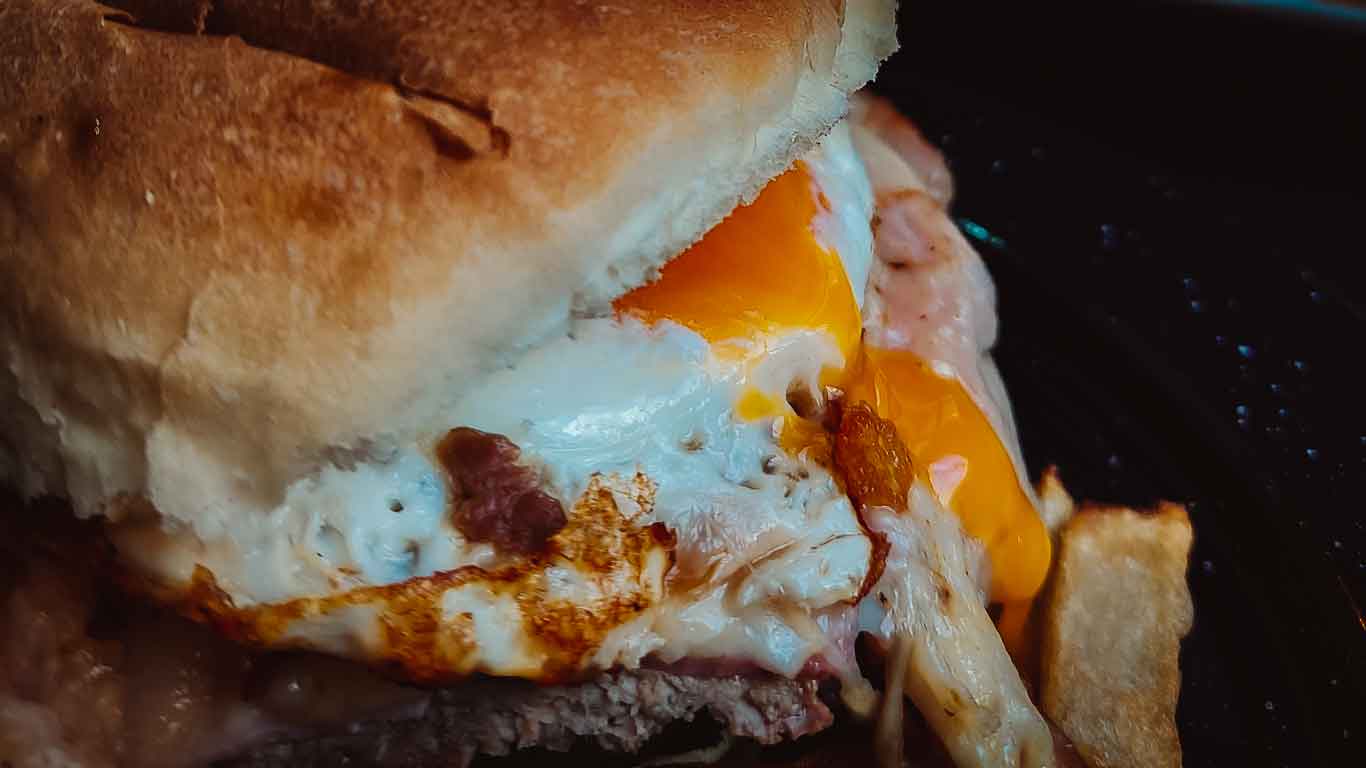 Imagen del típico bocadillo uruguayo, el chivito, donde la yema del huevo escurre delicadamente sobre los demás ingredientes, realzando el apetitoso conjunto de sabores.