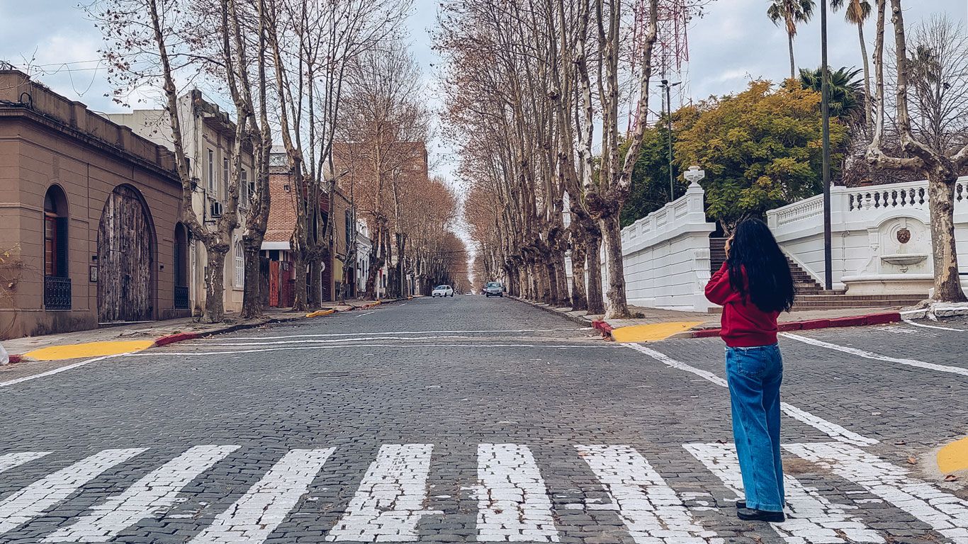Mujer en el Casco Histórico de Colonia del Sacramento, en Uruguay, tomando una foto de la calle empedrada con árboles sin hojas.