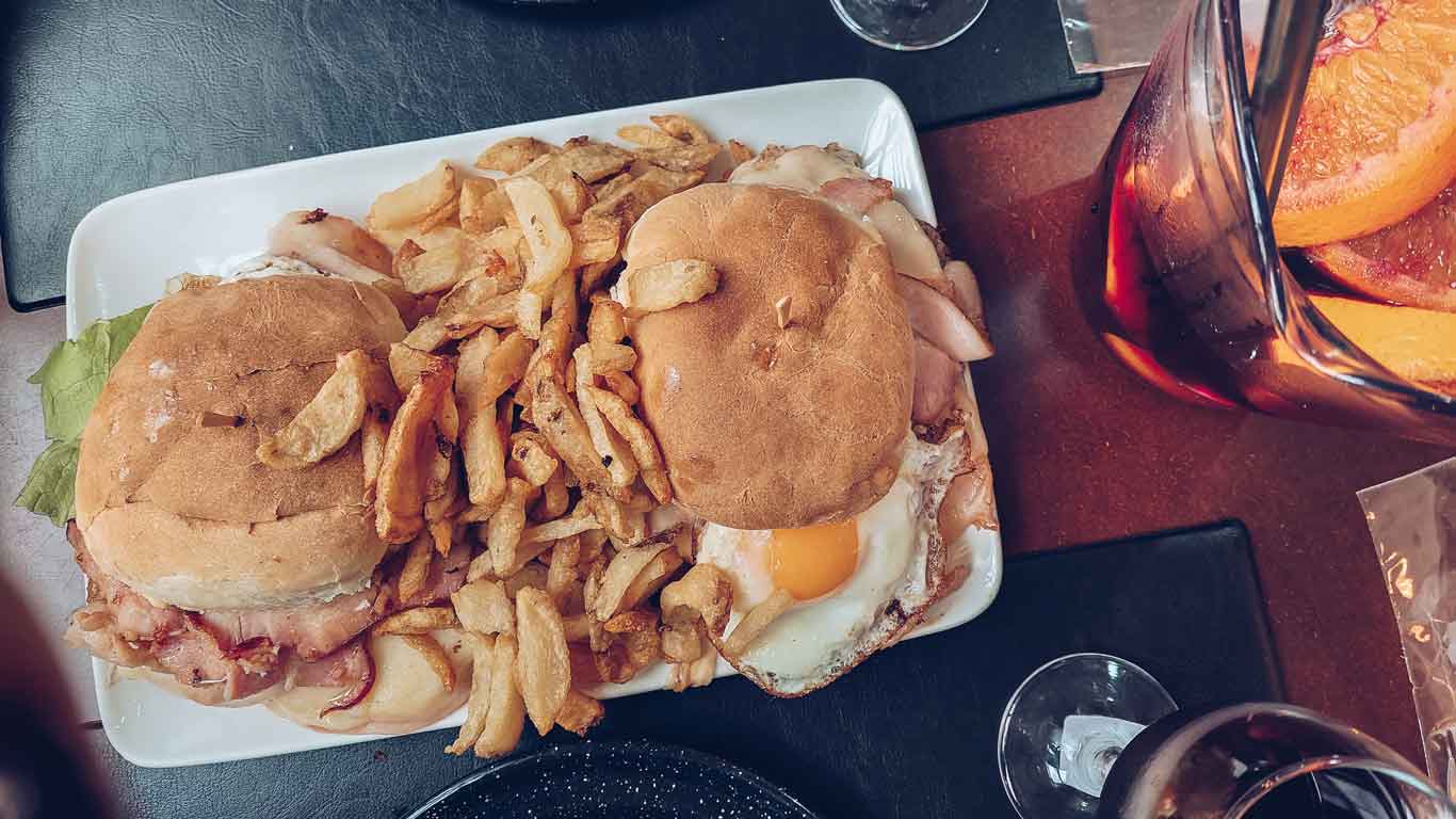 Foto de dos chivitos acompañados de patatas fritas, junto a una jarra de sangría.