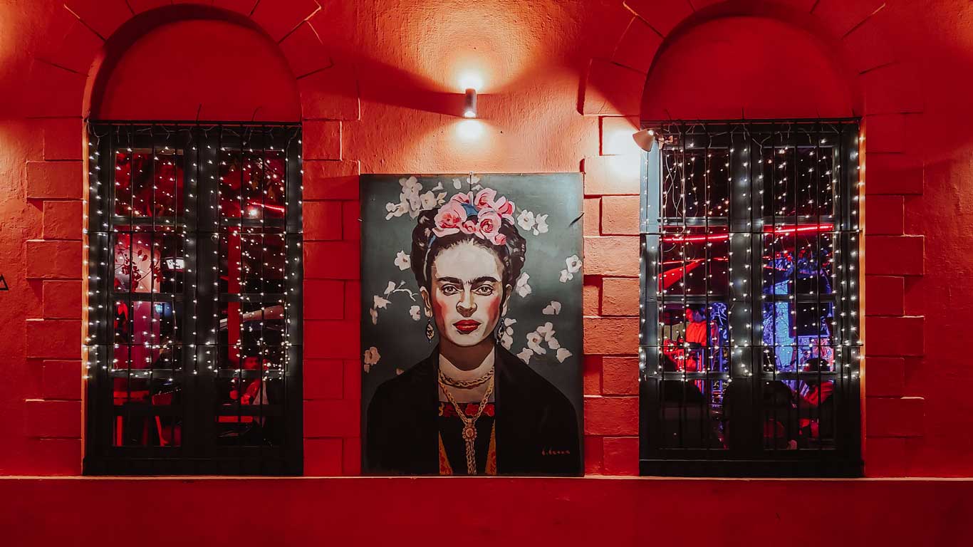 La imagen muestra la fachada del bar "La Locanda" en Colonia del Sacramento. La pared está pintada de rojo vibrante y adornada con una ilustración de Frida Kahlo, rodeada de flores blancas y con una corona de flores rosas en su cabeza. Dos ventanas arqueadas, protegidas por rejas y decoradas con luces de hada, permiten vislumbrar el interior acogedor del bar. Esta representación artística y el cálido ambiente iluminado reflejan el estilo único y la atmósfera acogedora de "La Locanda".