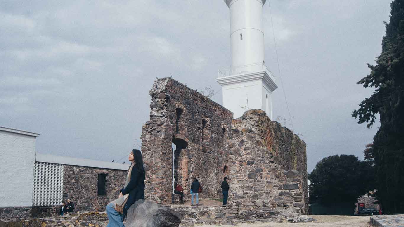 Personas explorando las ruinas históricas cerca del emblemático Faro de Colonia del Sacramento, bajo un cielo nublado, con una mujer en primer plano posando sobre una roca, reflejando la fusión de la historia y el turismo en Uruguay.