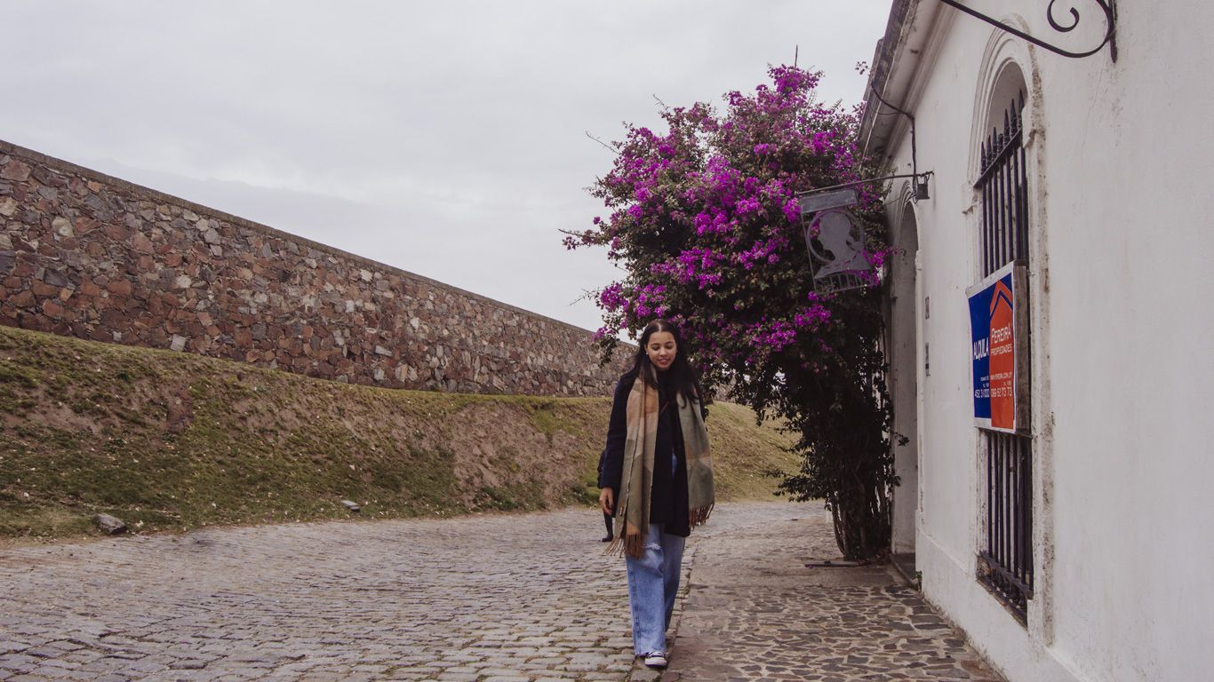 La imagen muestra a una joven caminando por una calle empedrada en Colonia del Sacramento. Ella pasa junto a un árbol de buganvilla con flores púrpura intensas, al lado de un edificio blanco. Al fondo, un muro de piedra rústico se extiende a lo largo del lado izquierdo de la imagen, añadiendo un ambiente histórico a la escena.