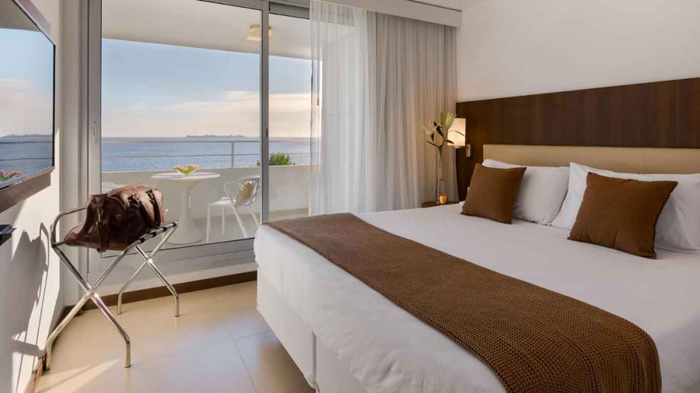 Una habitación moderna del  Dazzler by Wyndham con una amplia cama con sábanas blancas y cojines de acento marrones, una lámpara de noche, y un balcón con vistas al mar. Hay una bolsa de viaje de cuero marrón en un portaequipajes metálico, y una mesa redonda blanca con dos sillas en el balcón.