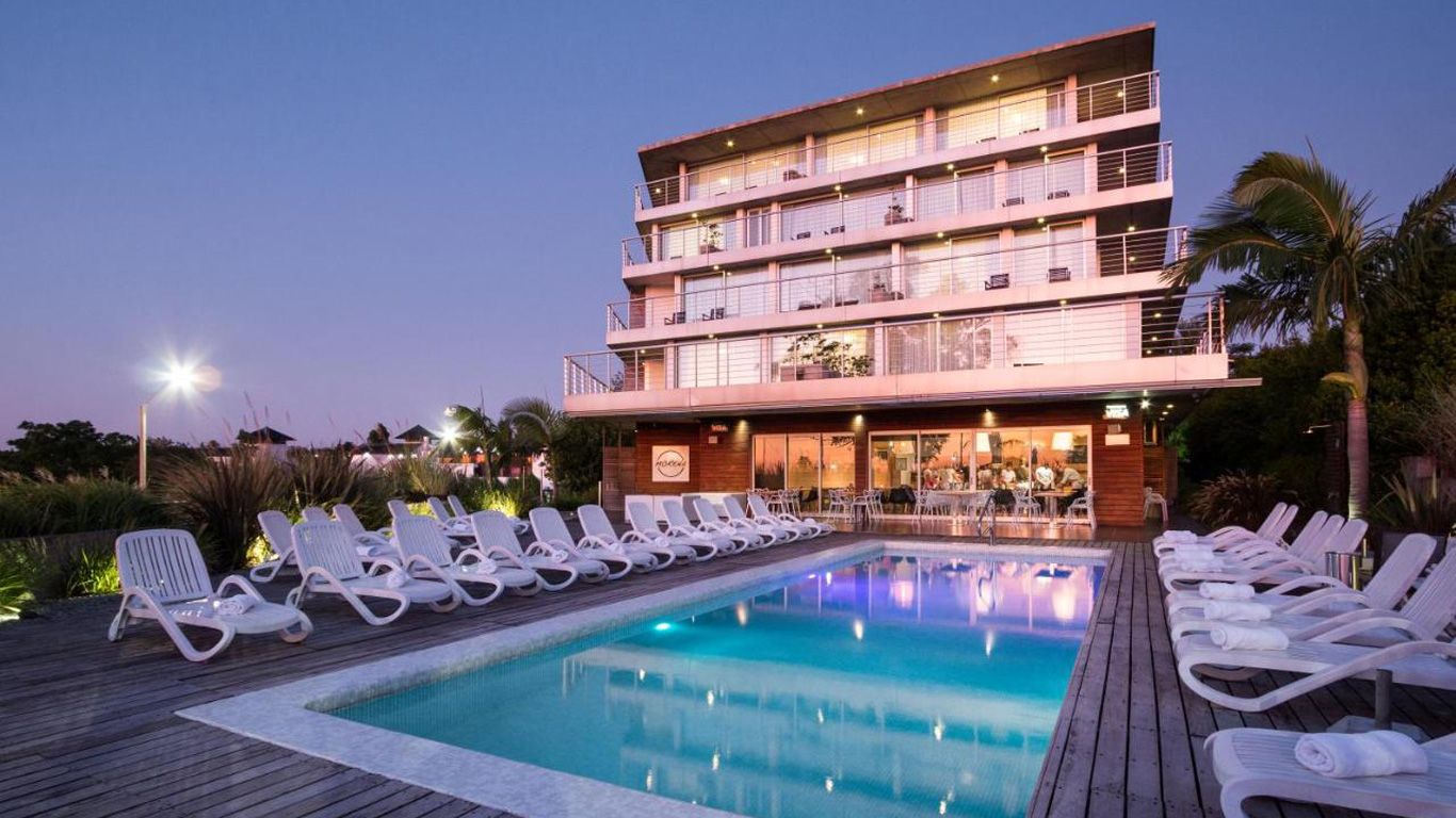 El crepúsculo cae sobre el Costa Colonia Boutique Hotel, con su moderna arquitectura de varios pisos que presenta balcones con vistas a una serena zona de piscina alineada con tumbonas blancas, acentuada por el suave resplandor de las luces junto a la piscina y un ambiente tranquilo.