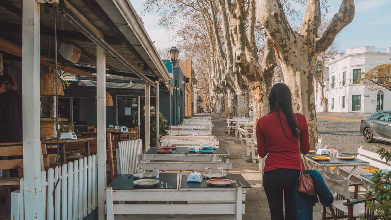 Una mujer caminando por la acera de Colonia del Sacramento, donde nos encontramos con numerosas sillas de un restaurante.