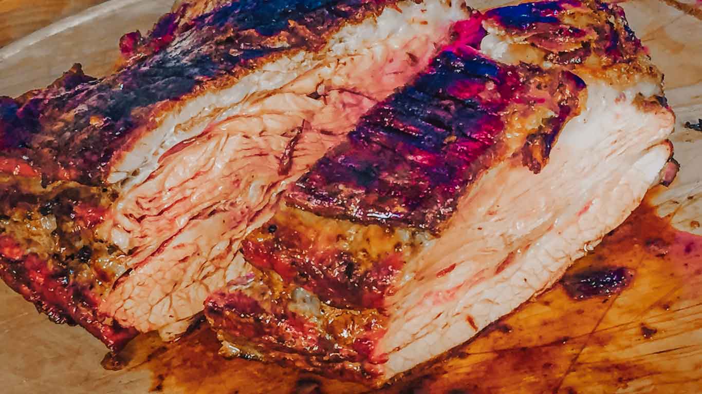 La imagen muestra un trozo de carne uruguaya recién asada, cortada y servida en una tabla de cortar de madera. La carne presenta un exterior perfectamente chamuscado con un interior jugoso y rosado, indicativo de una cocción a término medio. Los colores ricos y las texturas destacan la suculencia y la calidad típica de la barbacoa uruguaya, conocida localmente como "parrilla".