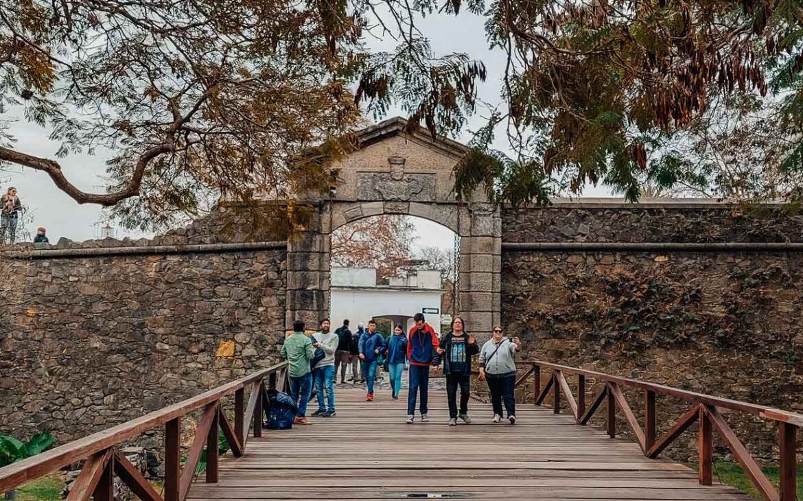 Visitantes cruzando un puente de madera hacia el histórico Portón de Campo en Colonia del Sacramento, Uruguay. El portón, hecho de piedra y con un arco en la parte superior adornado con un escudo, marca la entrada a la antigua fortaleza. Árboles y follaje decoran el entorno, añadiendo un toque natural a la escena histórica.