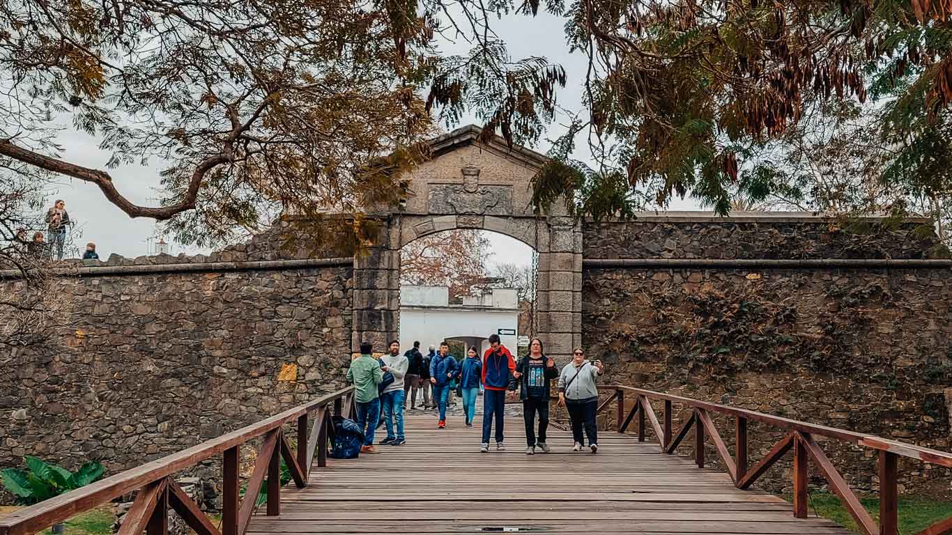 Visitantes cruzando un puente de madera hacia el histórico Portón de Campo en Colonia del Sacramento, Uruguay. El portón, hecho de piedra y con un arco en la parte superior adornado con un escudo, marca la entrada a la antigua fortaleza. Árboles y follaje decoran el entorno, añadiendo un toque natural a la escena histórica.