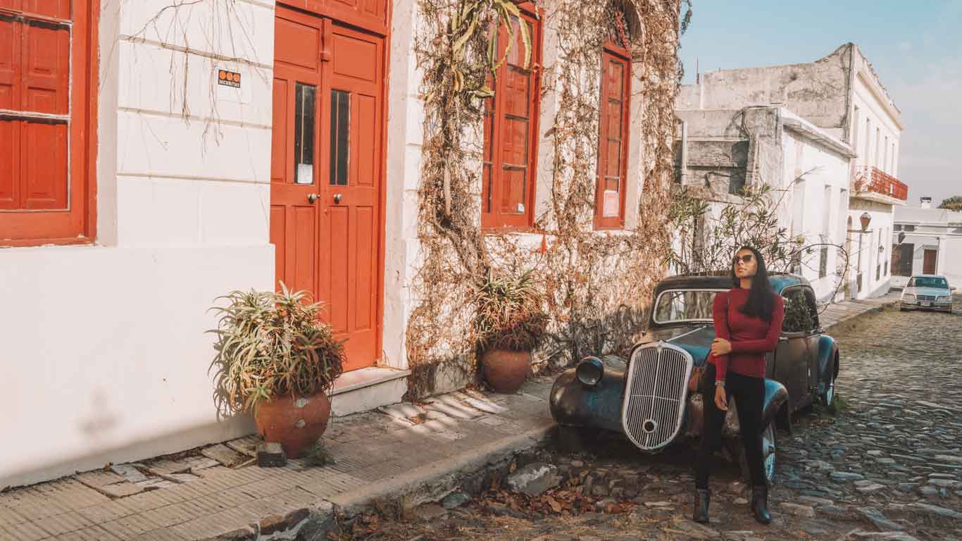 La imagen muestra una calle empedrada en Colonia del Sacramento, Uruguay, con una mujer posando junto a un coche antiguo estacionado frente a una casa con puertas y ventanas de color rojo. Las paredes de la casa están parcialmente cubiertas de enredaderas, añadiendo un toque pintoresco y nostálgico. La escena refleja el encanto histórico y colonial de la ciudad.