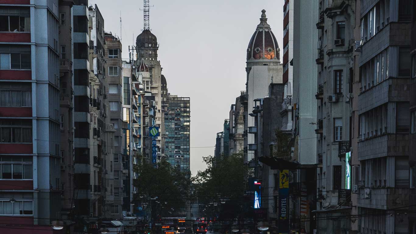 Vista de la Avenida 18 de Julio en Montevideo al atardecer, rodeada de altos y históricos edificios. En el horizonte, se destacan torres arquitectónicas, mientras las luces de la ciudad comienzan a brillar. Este es un excelente lugar donde alojarse en Montevideo.