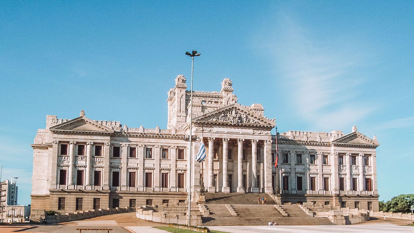 Fachada del Palacio Legislativo de Montevideo, un imponente edificio neoclásico con columnas y esculturas detalladas. La bandera de Uruguay está ondeando frente al palacio, que es un importante símbolo político e histórico del país.