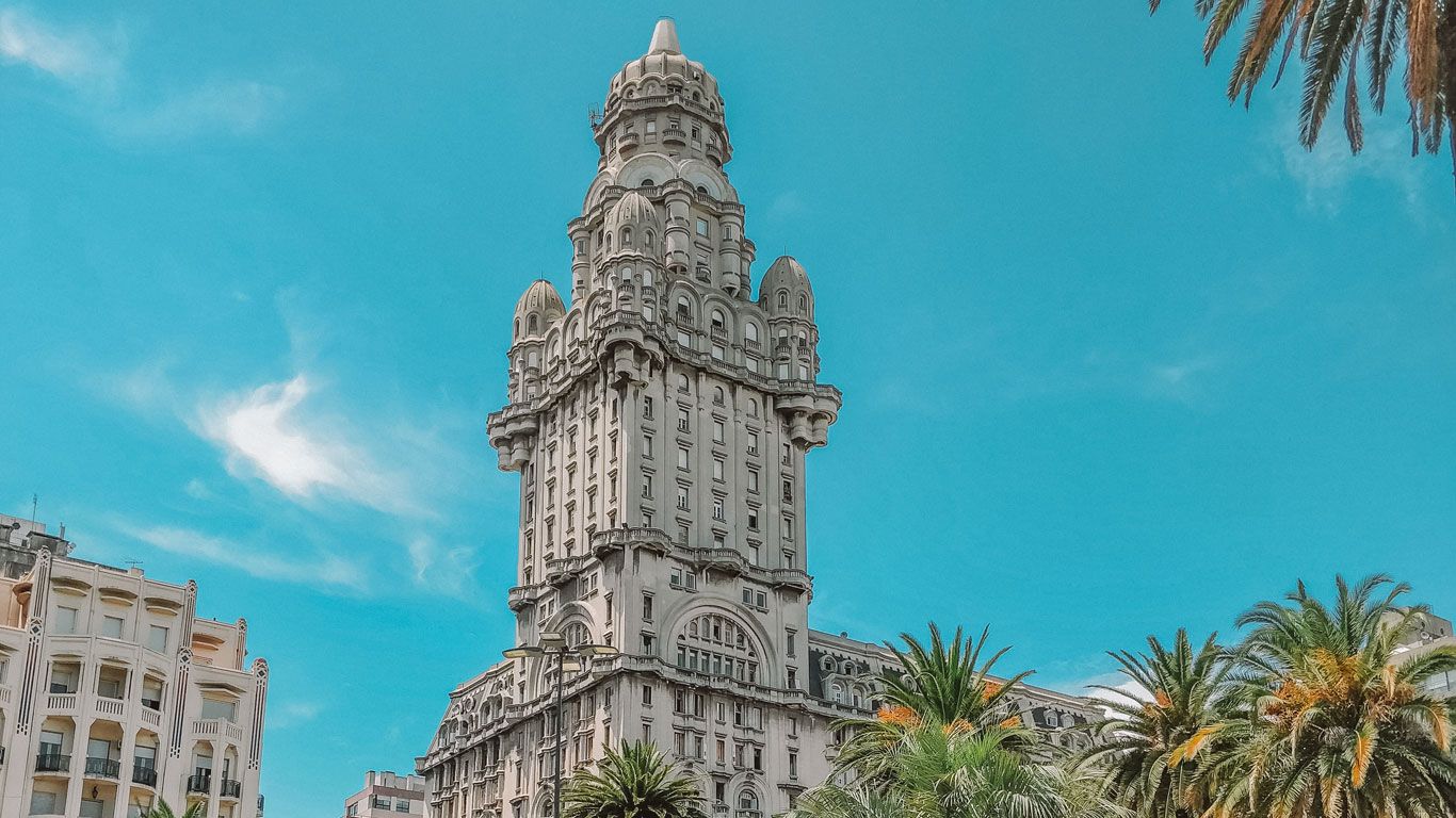 La imagen muestra el imponente Palacio Salvo en Montevideo, una icónica construcción que se destaca en el paisaje urbano de la ciudad. Este edificio, que fue el más alto de América del Sur en su tiempo, exhibe una arquitectura ecléctica con influencias góticas y neorrenacentistas. Su torre central se eleva majestuosamente, adornada con detalles ornamentales y coronada por una cúpula, rodeada de palmeras que aportan un toque tropical al entorno urbano.