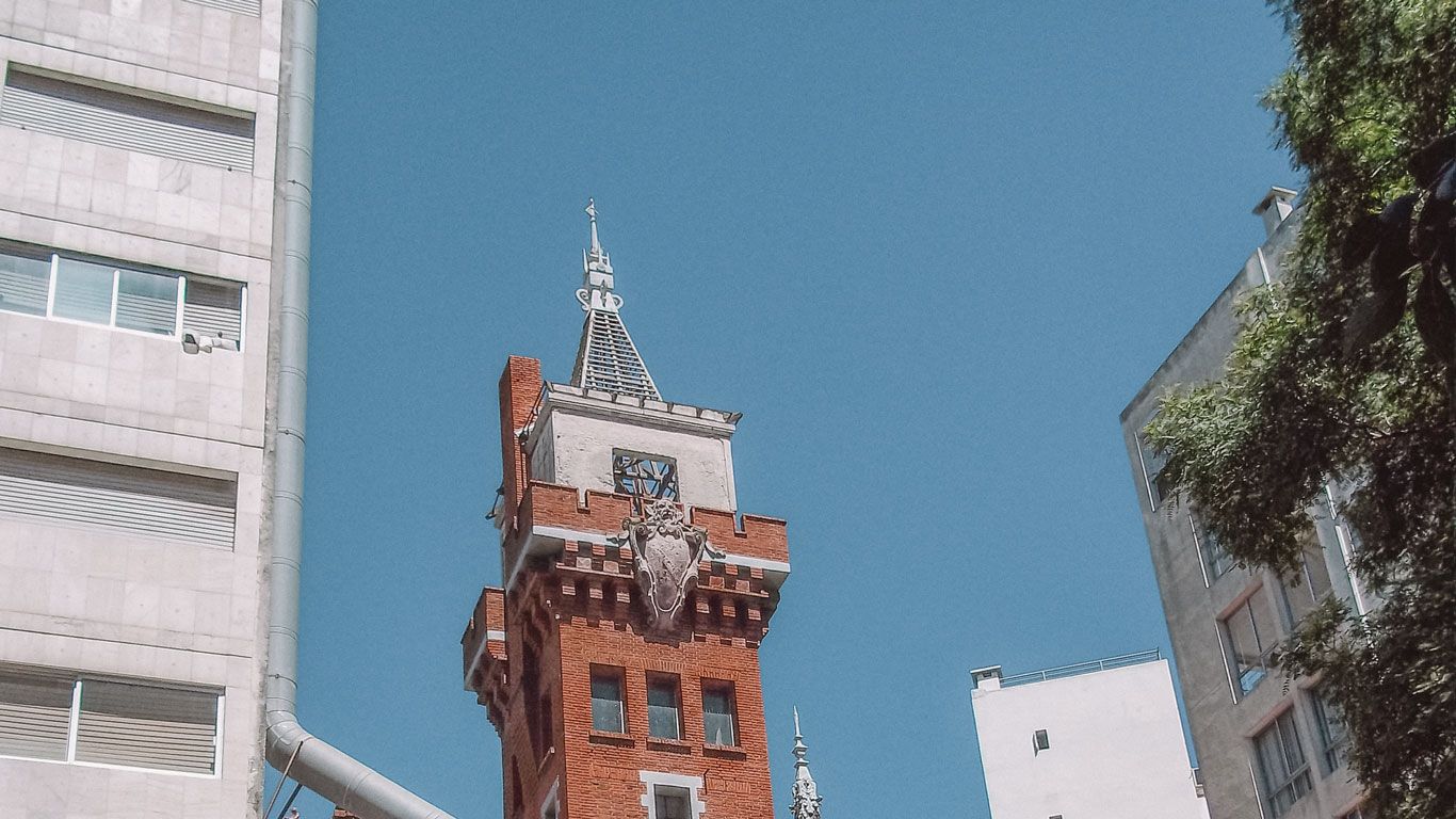 Parte superior del Castillo Pittamiglio en Montevideo, destacando su arquitectura única con ladrillos rojos y detalles ornamentados. El edificio está rodeado por construcciones modernas, creando un contraste interesante.