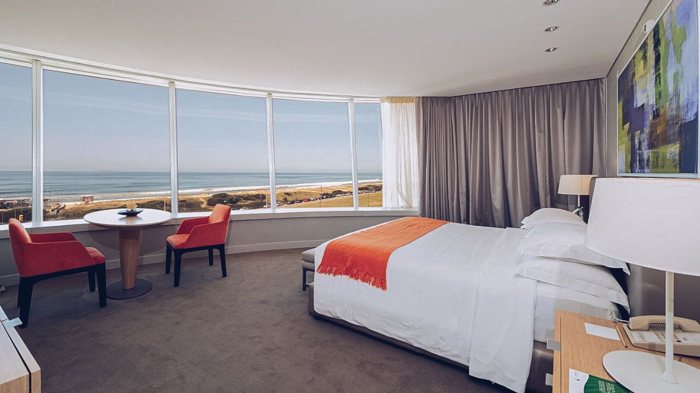 La imagen presenta una habitación del The Grand Hotel en Punta del Este, destacando por su elegante diseño interior y una panorámica vista al mar. Desde una amplia ventana curva, se observa la playa y el horizonte oceánico, proporcionando un escenario relajante y luminoso. La habitación está amueblada con una cama grande con sábanas blancas y una manta naranja, acompañada de sillas rojas en una mesa redonda.