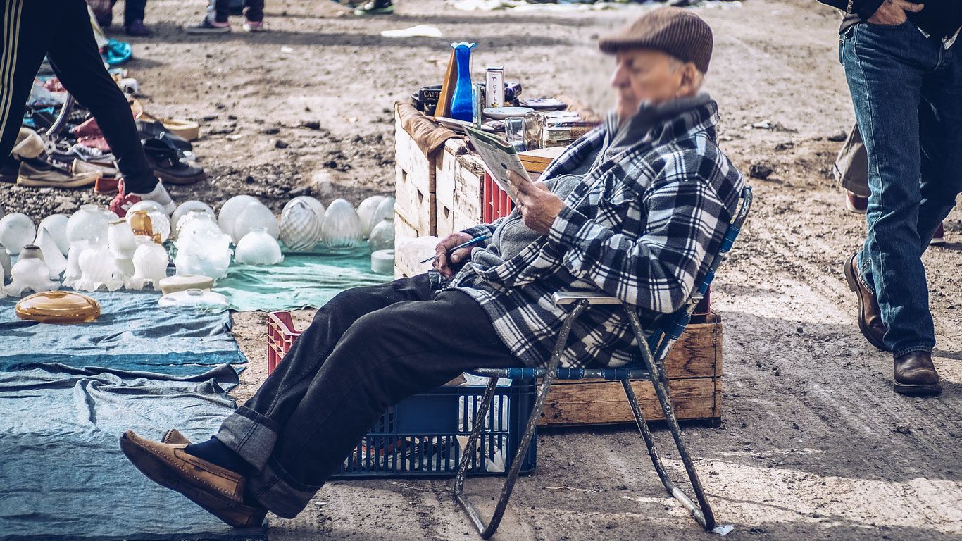 Hombre mayor sentado en una silla en la Feria Tristán Narvaja en Montevideo, leyendo un periódico. Alrededor, hay varios artículos usados a la venta, incluyendo lámparas y objetos de vidrio. La feria es conocida por su diversidad de productos y ambiente acogedor.