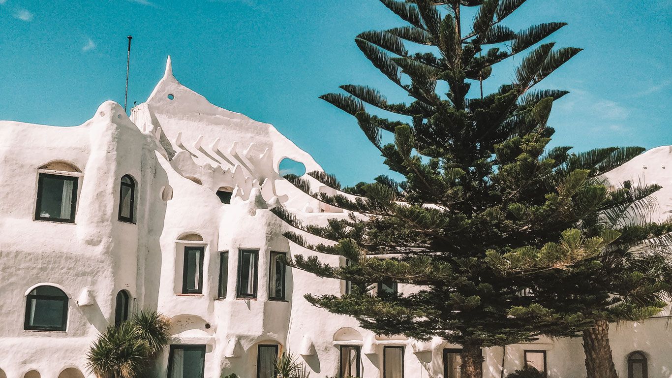 La imagen muestra la emblemática Casapueblo, situada en Punta del Este, un edificio que se distingue por su arquitectura única que emula formas orgánicas y escultóricas. Esta construcción blanca, que parece esculpida en la roca, contrasta con el cielo azul claro y está rodeada de vegetación típica del litoral, incluyendo un pino prominente en primer plano.