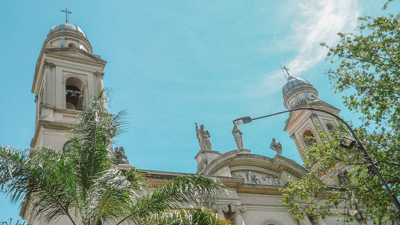 Fachada de la Catedral Metropolitana de Montevideo, con sus torres gemelas y estatuas decorativas en la parte superior. La catedral está rodeada de palmeras y árboles, bajo un cielo azul claro.