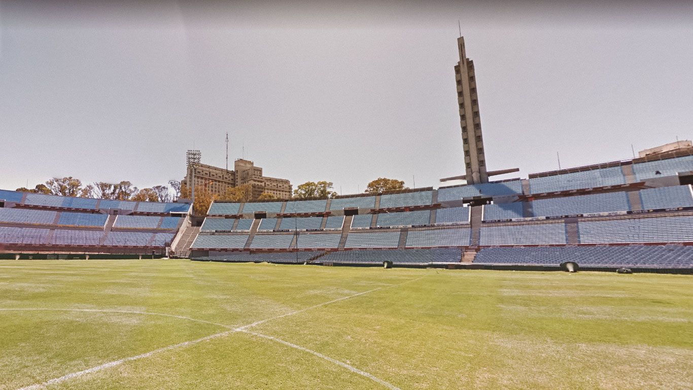 Vista interior del Estadio Centenario en Montevideo, con gradas vacías y césped verde bien cuidado. Se destaca la torre de los homenajes al fondo.