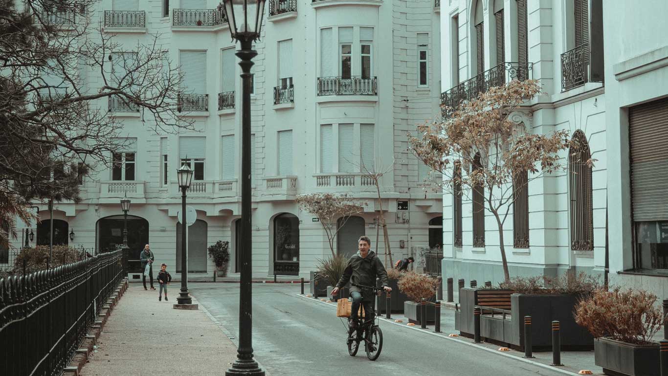 La imagen muestra una calle tranquila en Montevideo, Uruguay, con un hombre montando una bicicleta y un niño corriendo al lado de edificios antiguos y elegantes. Los árboles sin hojas y el ambiente sereno sugieren que es otoño o invierno. La arquitectura y las farolas de estilo clásico destacan el encanto histórico de la ciudad.