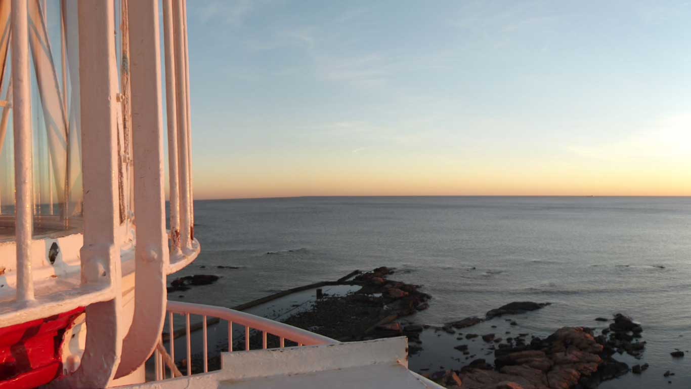 Vista del Faro de Punta Carretas en Montevideo al atardecer, con la estructura blanca del faro en primer plano y el mar tranquilo al fondo. El cielo está teñido de tonos de naranja y azul, creando una atmósfera serena y pintoresca.