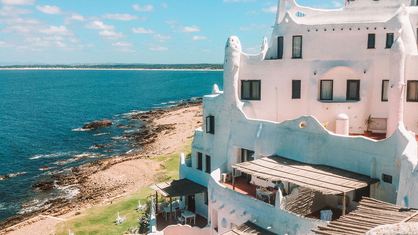 La imagen muestra Casapueblo, un famoso edificio blanco con forma de escultura ubicado a lo largo de la costa rocosa de Punta del Este, Uruguay. El edificio, con su estilo arquitectónico único, sus terrazas y su blanco puro contra el azul del mar y el cielo, ofrece una escena cautivadora.