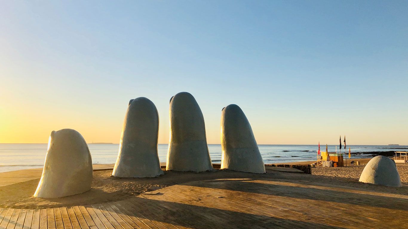 La icónica escultura La Mano del artista chileno Mario Irarrázabal, que representa cinco dedos gigantes emergiendo de la arena, en la Playa Brava de Punta del Este, Uruguay, durante un tranquilo amanecer con cielo despejado.